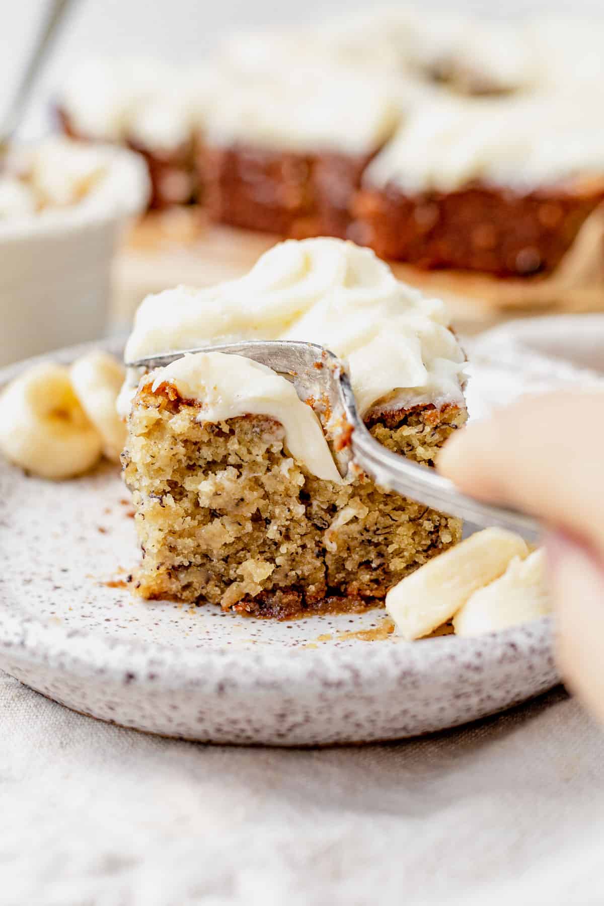 a fork cutting into a piece of banana cake with cream cheese frosting