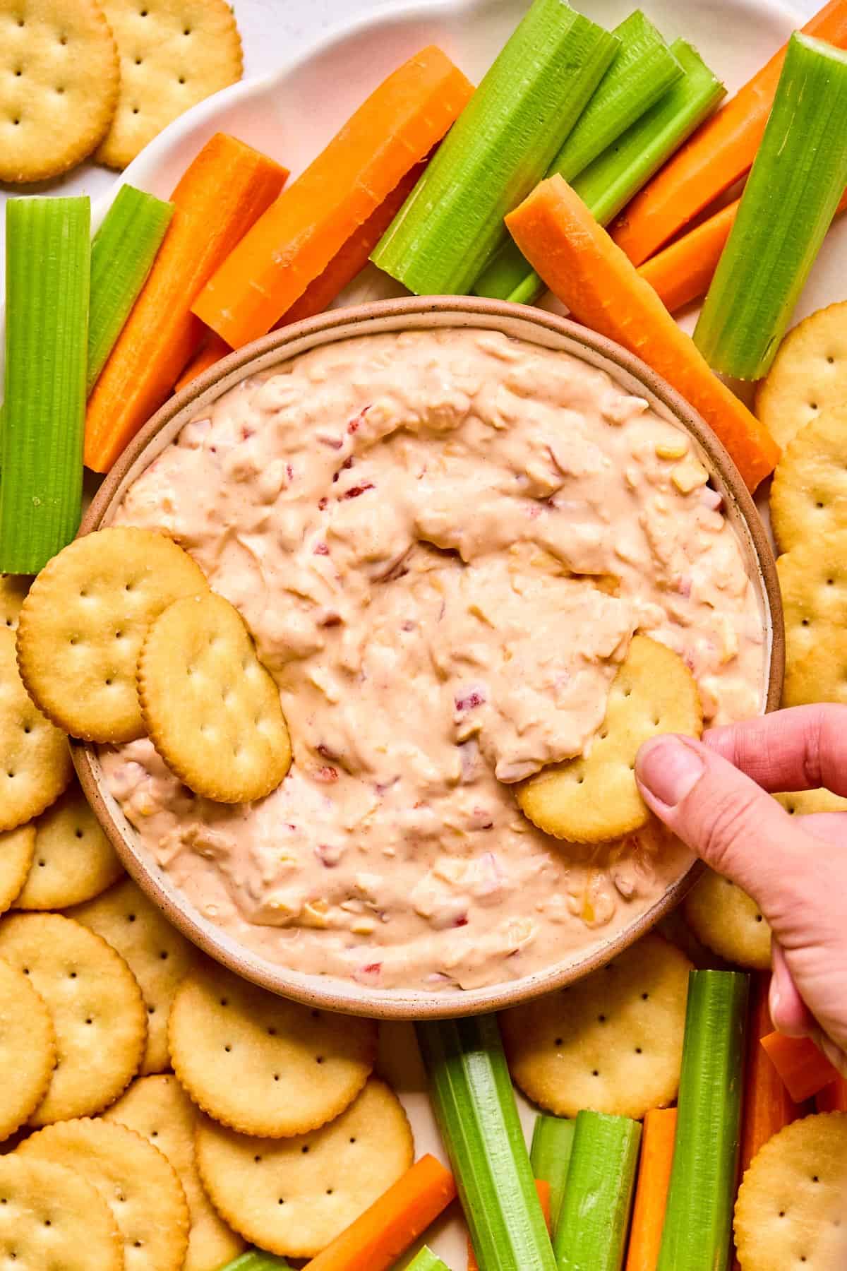 hand reaching with a ritz cracker to dip into pimento cheese dip surrounded by carrots, celery, and crackers.