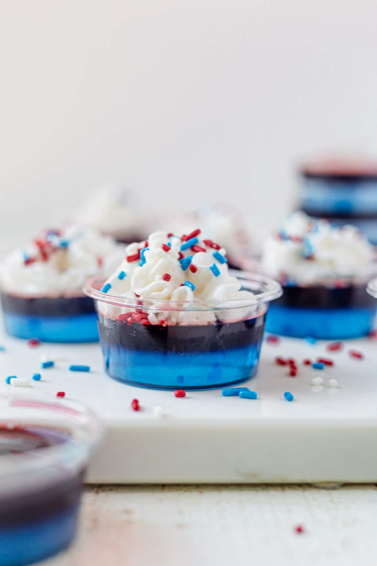 red white and blue jello shots on a white tray topped with whip cream and red and blue sprinkles