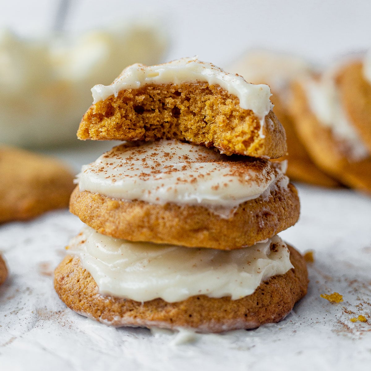 Three pumpkin cookies with cream cheese frosting are stacked, topped with a generous layer of white icing and sprinkled with spice. The top cookie is broken in half, revealing a soft, moist inside. Other cookies are blurred in the background.