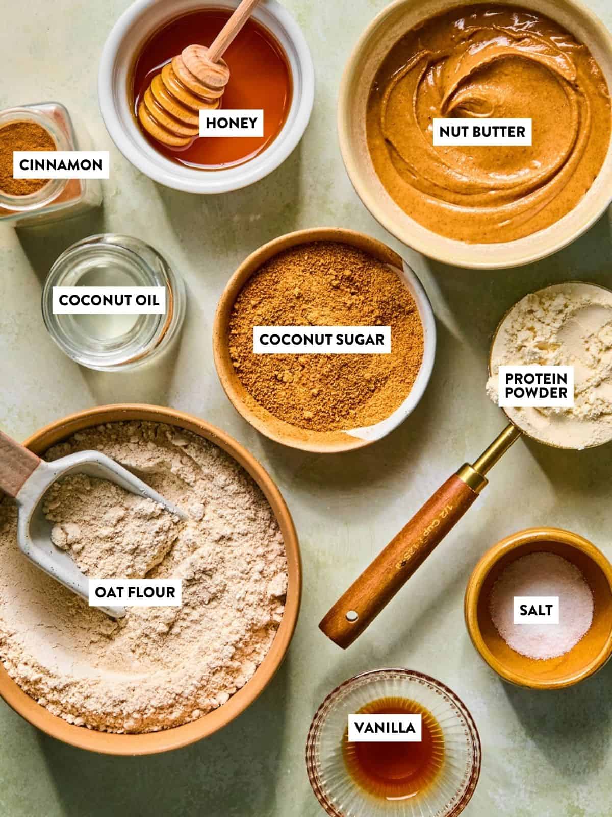 Various baking ingredients in bowls and jars, including labeled cinnamon, honey, nut butter, coconut oil, coconut sugar, protein powder, oat flour, salt, and vanilla—everything you need to make delicious snickerdoodle protein balls—arranged on a light surface.