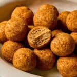 A close-up of a pile of round, golden snickerdoodle protein balls on a white plate, with one ball showing a bite taken out to reveal a dense, light brown interior.