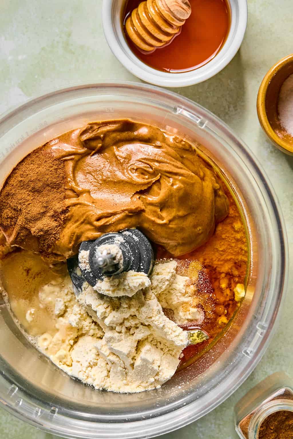 A food processor bowl filled with almond butter, almond flour, cinnamon, honey, and vanilla—for making snickerdoodle protein balls—with honey and salt in small dishes nearby.