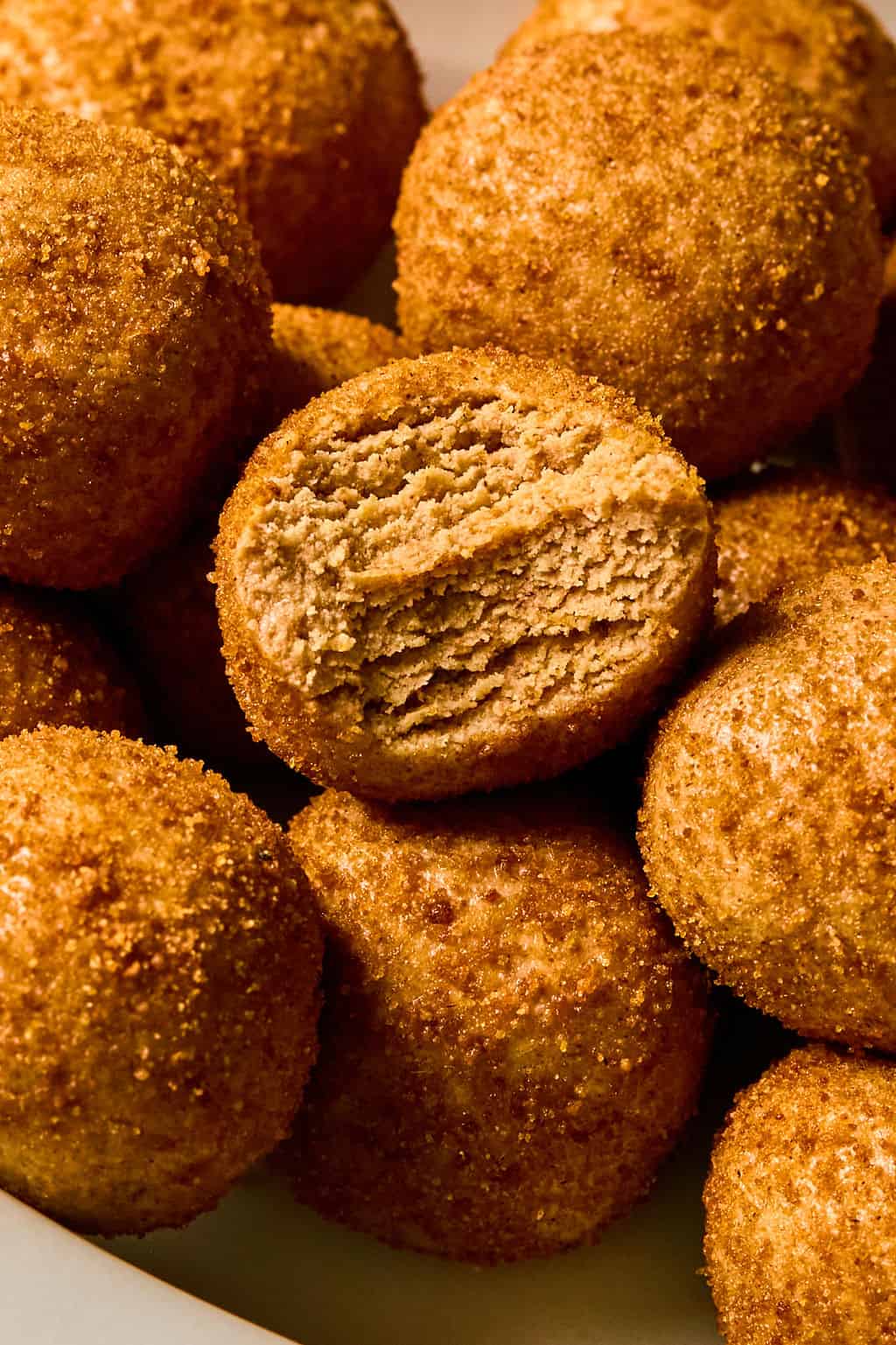 A close-up of golden, round, snickerdoodle protein bites, one with a bite taken out to reveal a soft, layered interior. The balls are piled together on a white surface.