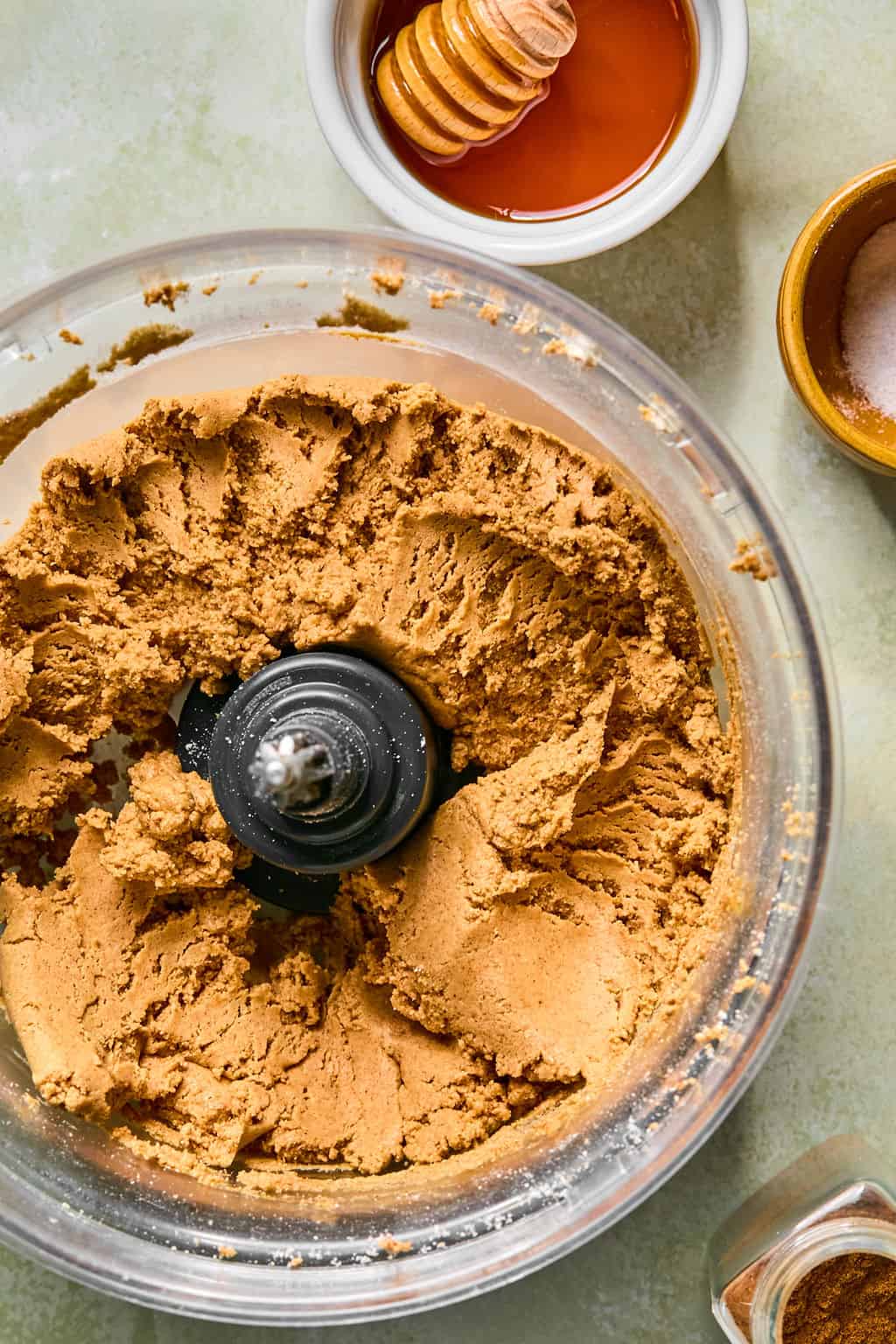A food processor with blended snickerdoodle protein ball dough, sits on a counter. Nearby are a small bowl of honey with a dipper, a bowl of salt, and a jar of cinnamon.