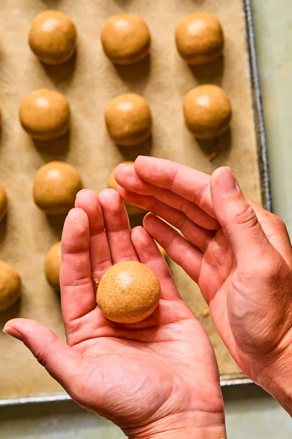Hands shaping snickerdoodle protein ball dough into a ball above a baking sheet lined with parchment paper, with several evenly spaced snickerdoodle protein balls already placed on the tray, ready for baking.