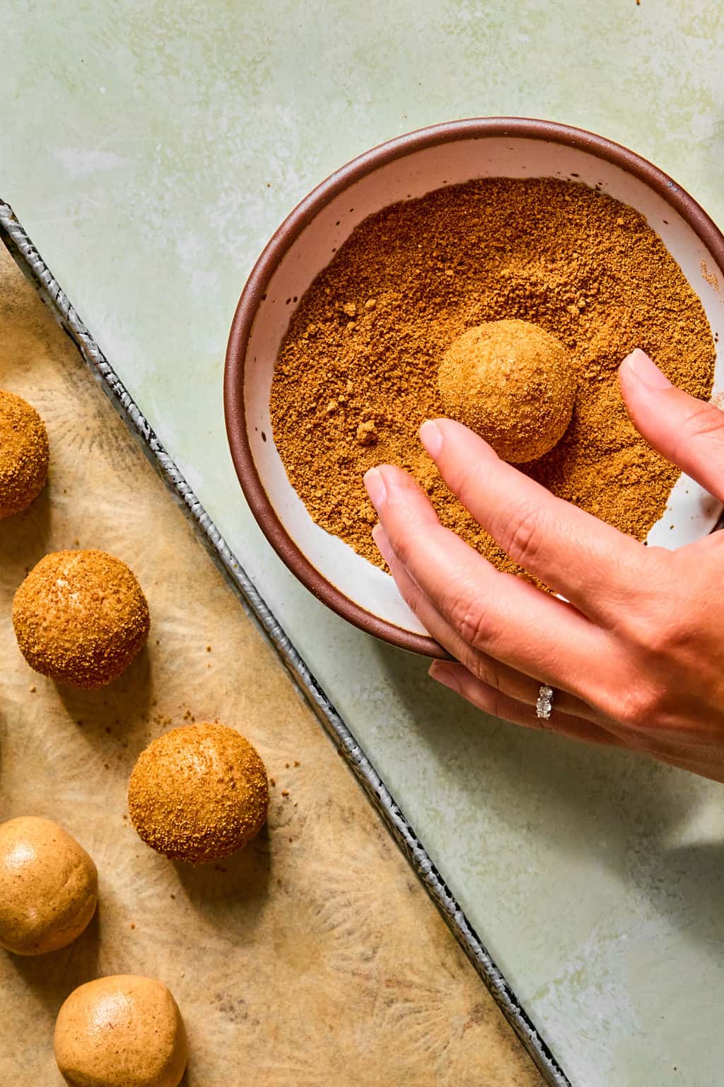 A hand rolls a dough ball in a bowl of brown crumbs to make snickerdoodle protein balls, with more coated balls resting on a parchment-lined baking tray nearby.