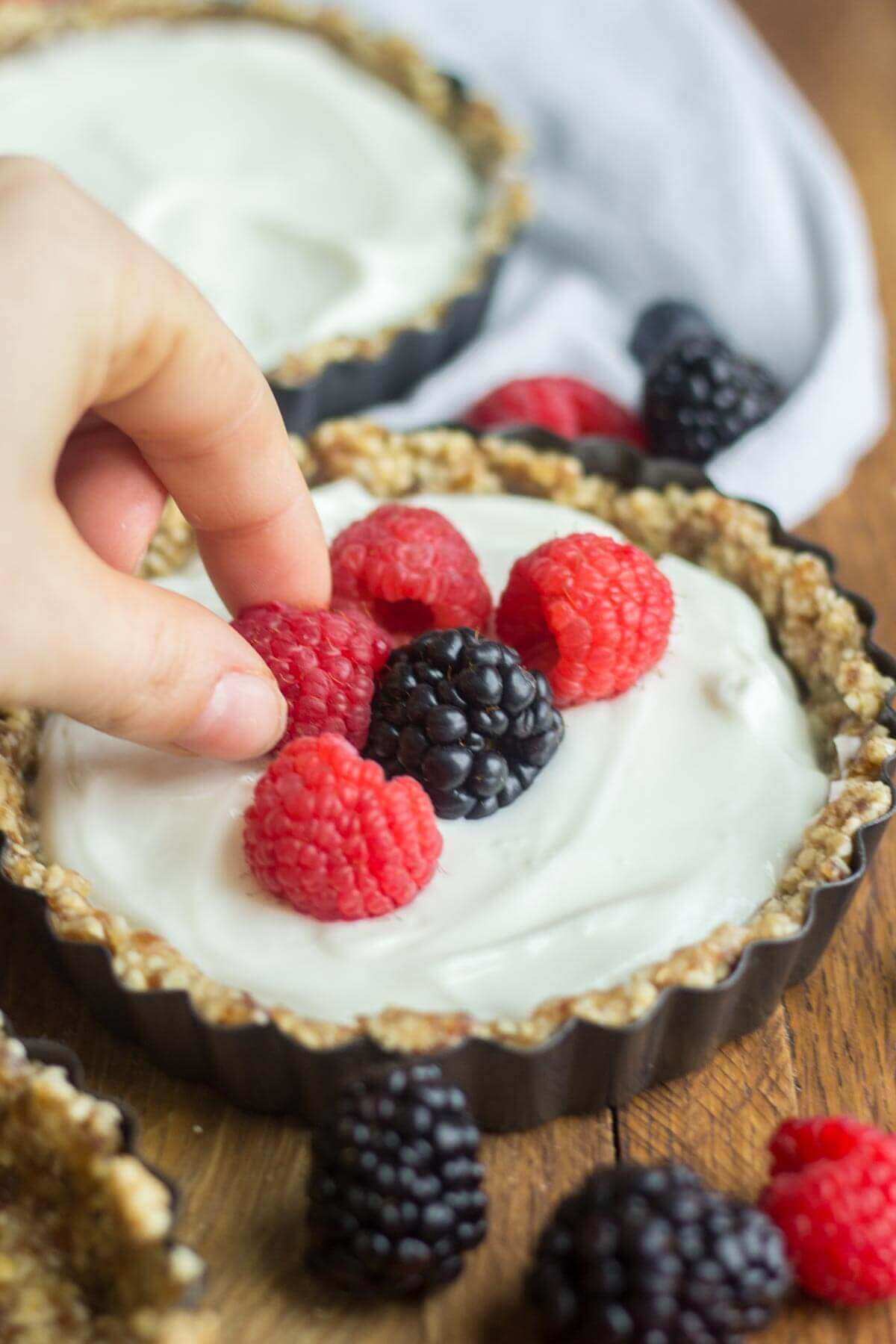 hand placing raspberry on top of mini greek yogurt fruit tart filled with fresh berries