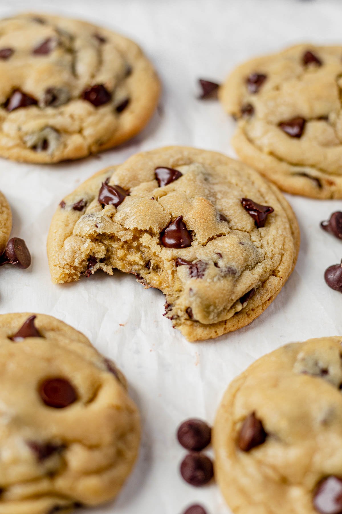 six freshly baked chocolate chip pudding cookies on a piece of parchment paper with melted chocolate chips on top and a bit taken out of the center one so you can see the gooey inside