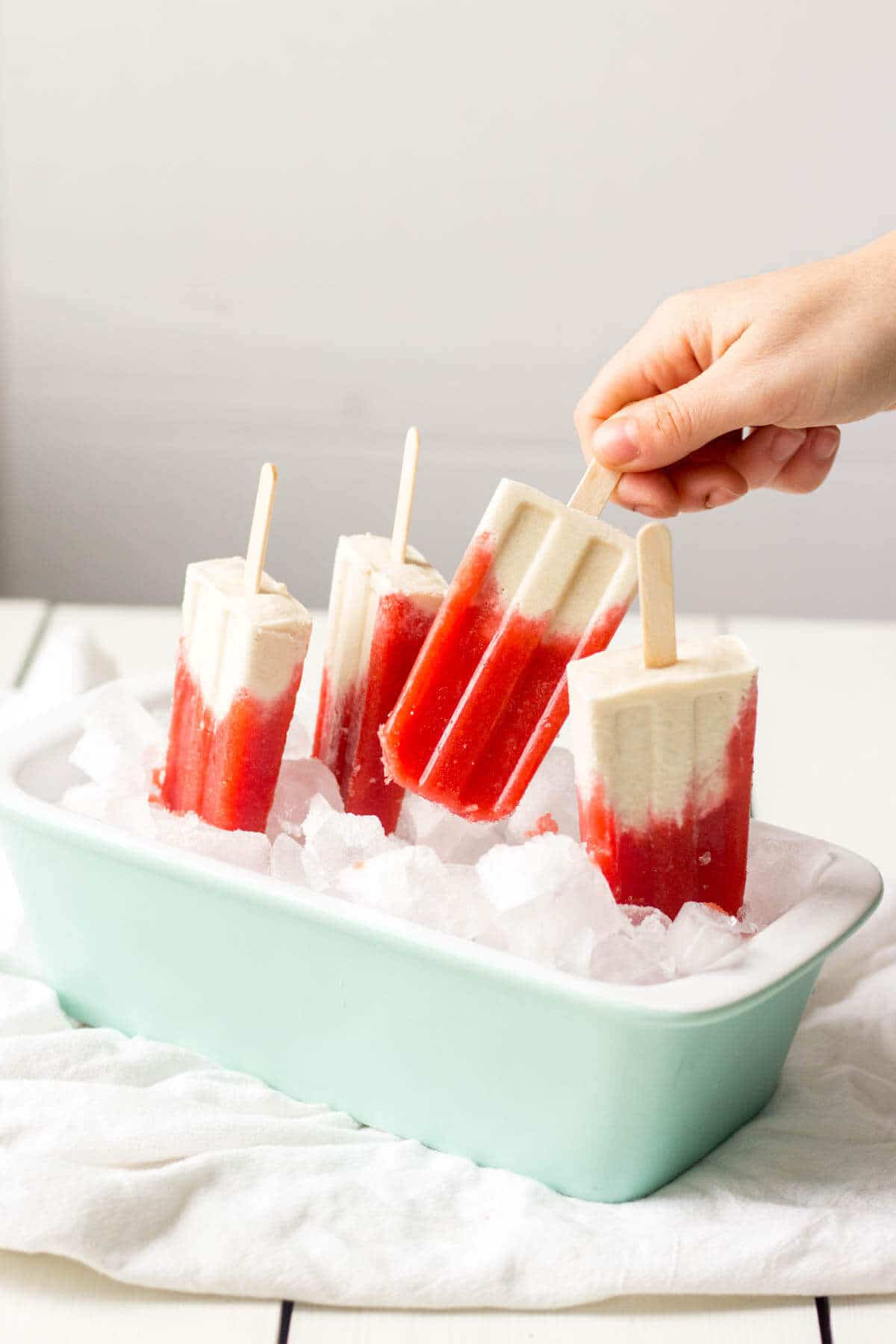 a hand reaching to grab the popsicle stick of a Strawberries and Cream Vegan Popsicle that is standing upsaide down in a container of ice