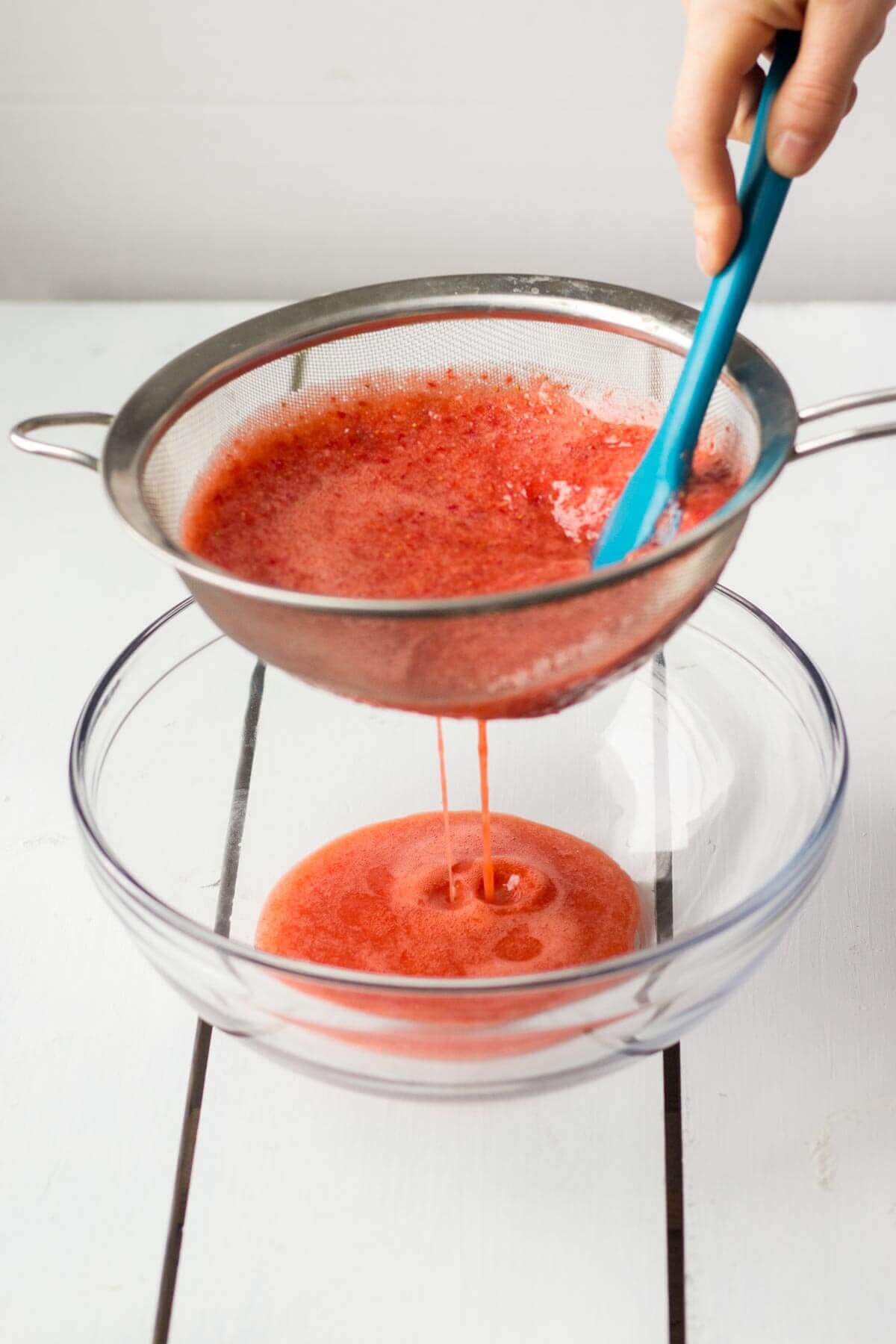 blue spatula pressing blended strawberries up against a strainer as it strains the strawberries into a glass bowl