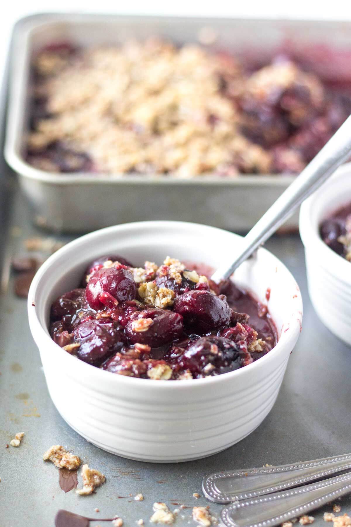 A silver spoon dipped into a white ramuken with easy cherry crisp recipe, cherries and dark chocolate and topped with a gluten free oat crust. 