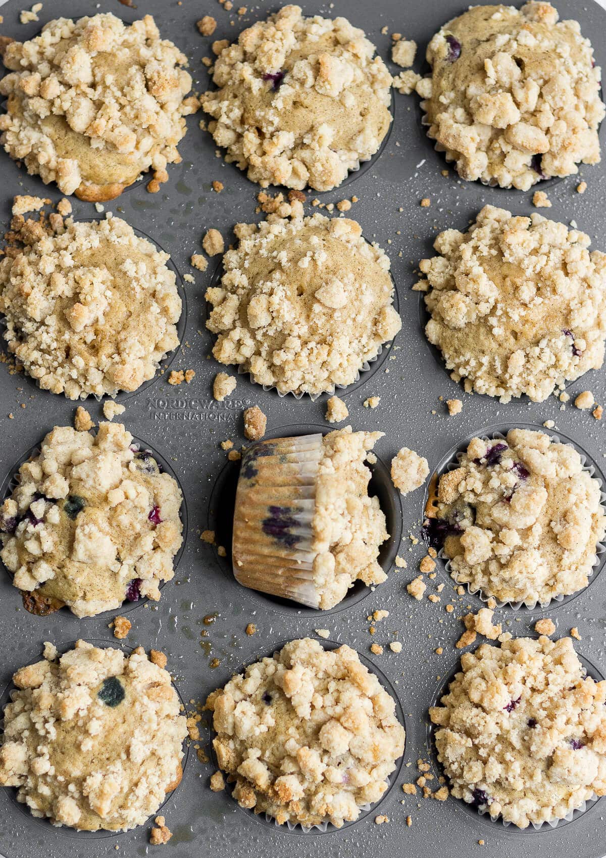 blueberry muffins in the muffin tins with crumb topping covering the tops