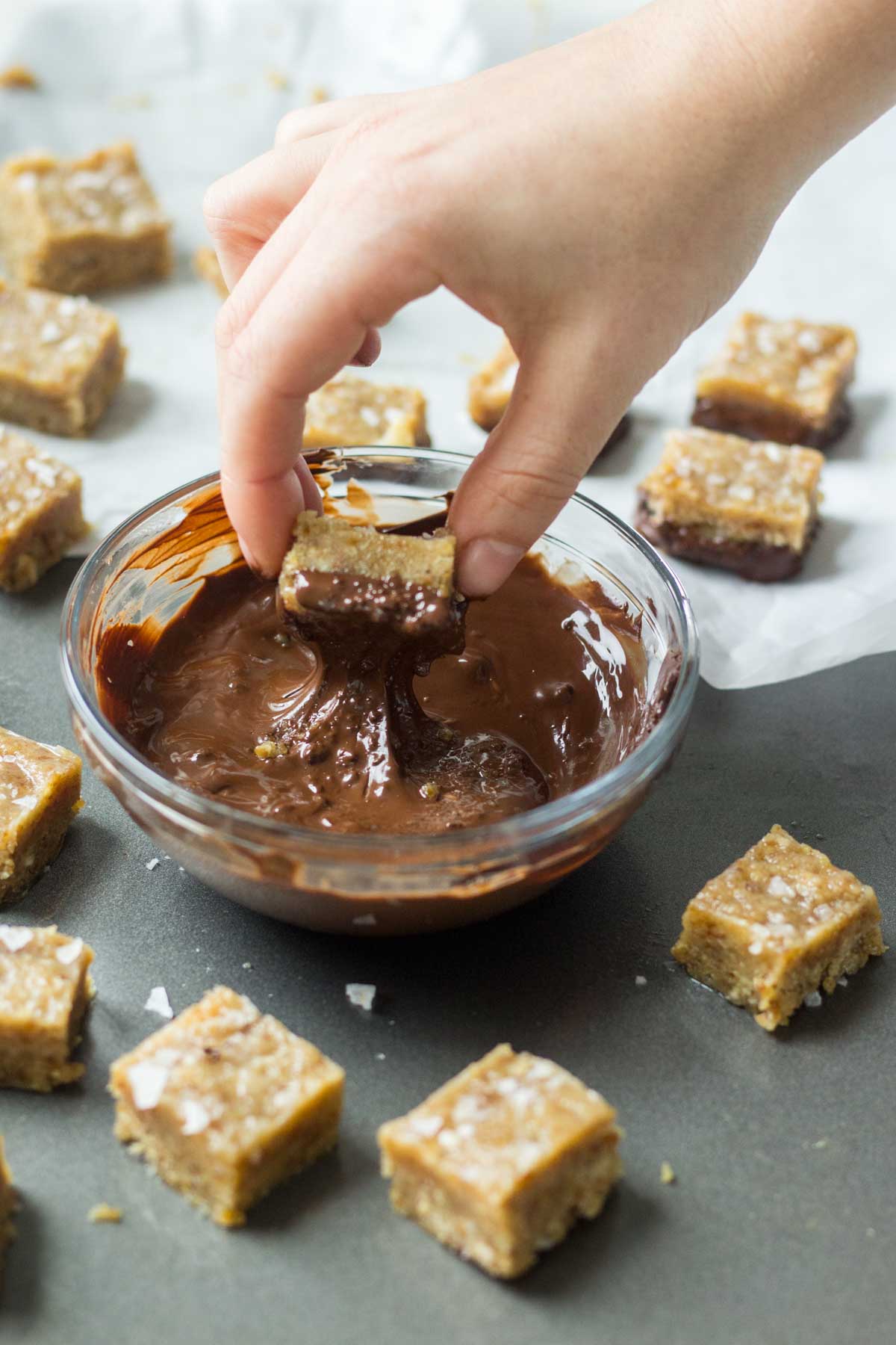 hand dipping healthy salted caramel into a bowl of melted chocolate and other pieces of salted caramel laying on the cookie sheet next to it