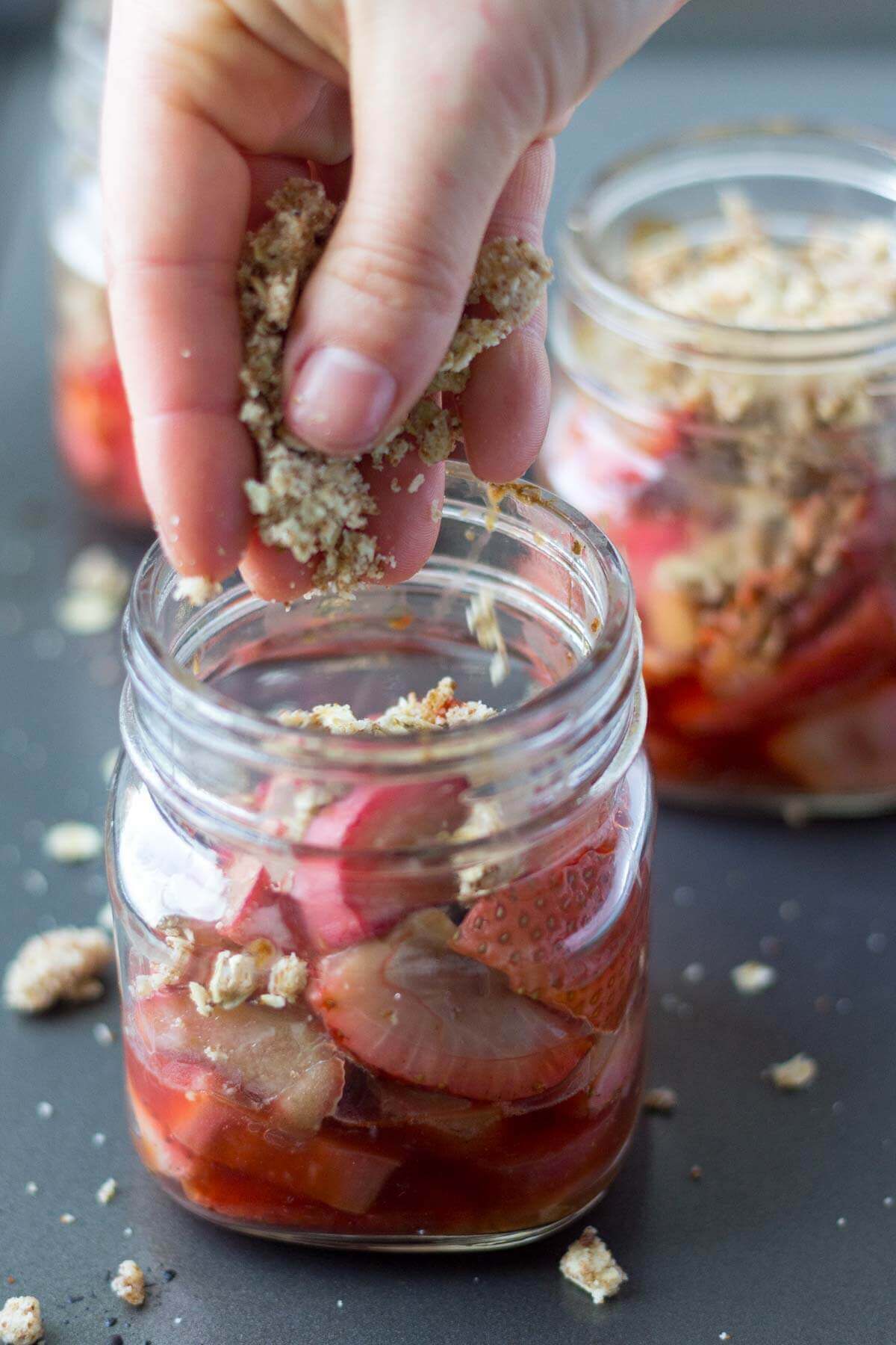 a hand dropping granola into a glass jar of strawberry rhubarb crisp