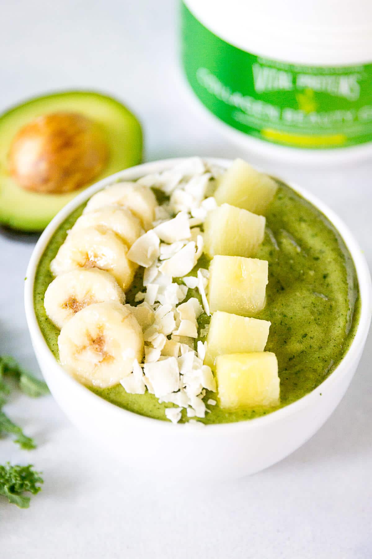 green smoothie bowl in a white bowl, topped with banana slices, coconut flakes, and pineapple chunks and next to a half an avocado