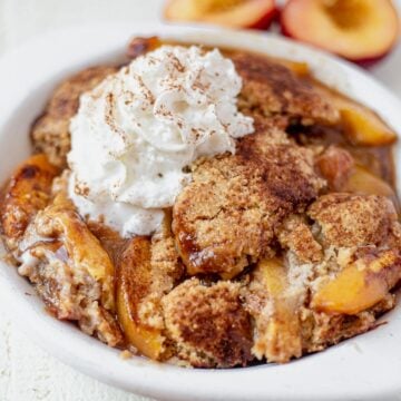 a large serving of gluten free peach cobbler in a white dish topped with whipped cream and cinnamon