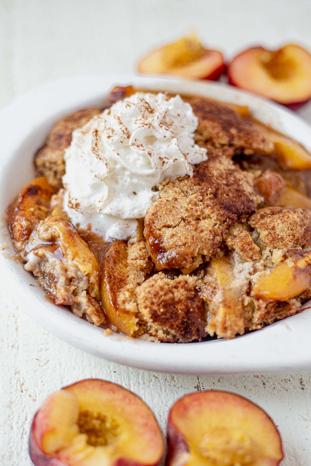 gooey gluten free peach cobbler in a white baking dish topped with fresh whipped cream and cinnamon