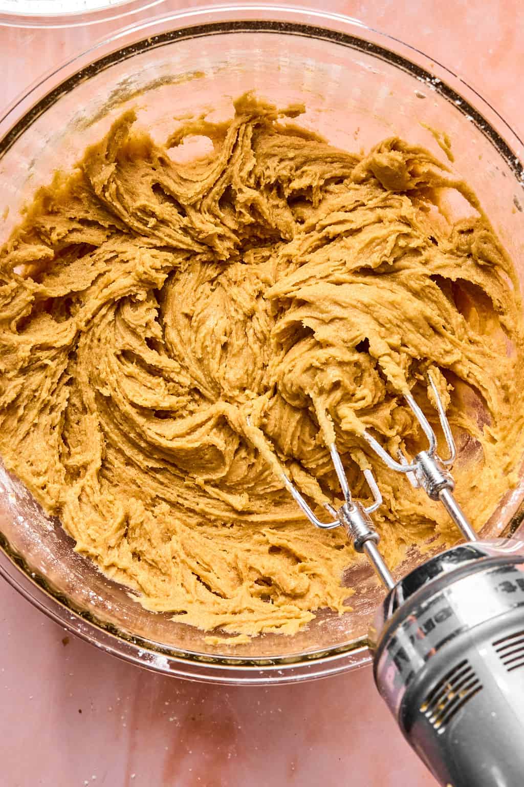 An electric hand mixer blending light brown apple pie bars cookie dough in a glass bowl on a pink countertop.