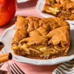A close-up of a square slice of apple pie bar on a white plate, showing layers of spiced apples and golden-brown crust. Cinnamon sticks, a fork, and an apple are nearby on a pink surface, capturing the classic charm of homemade apple pie bars.