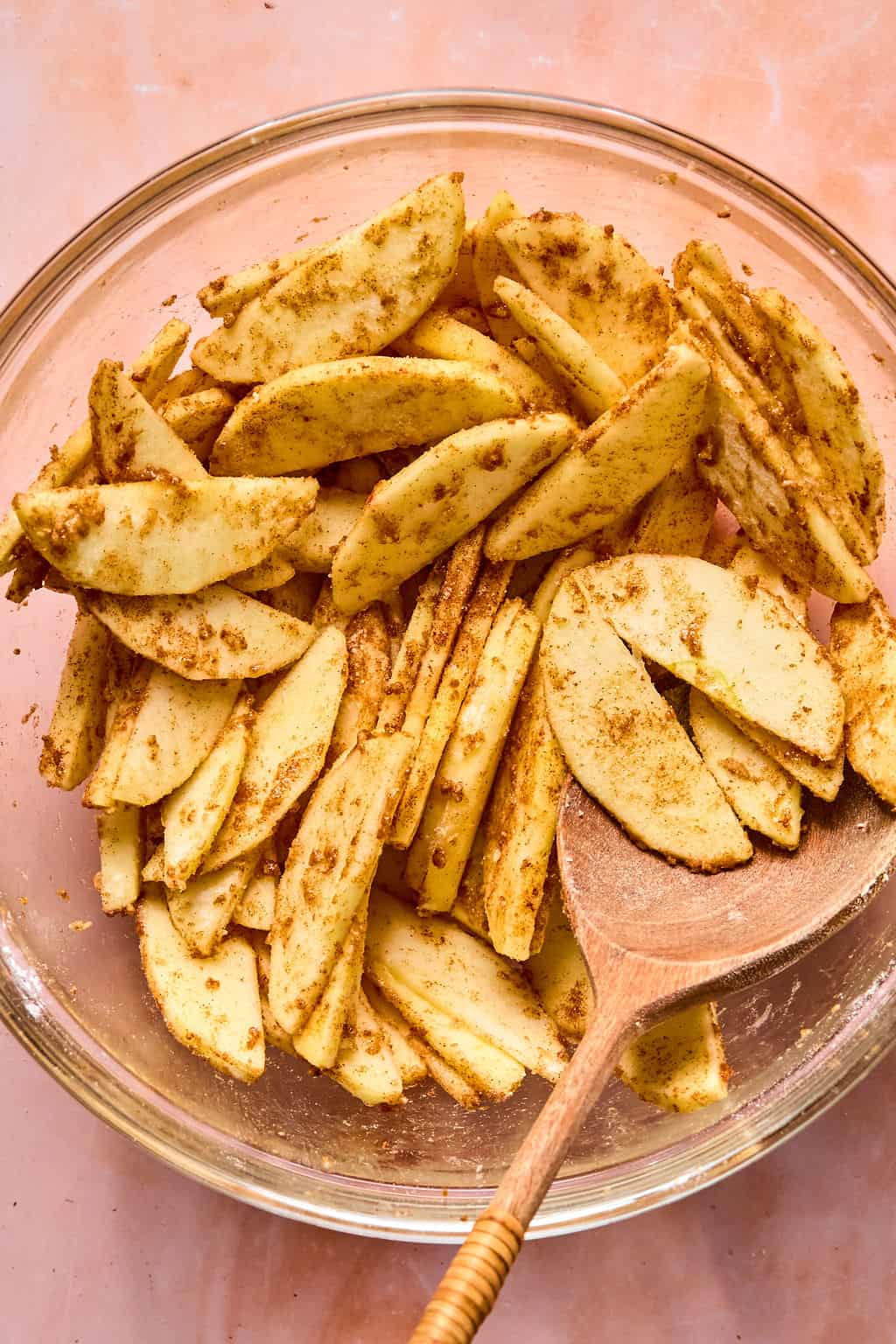 Sliced apples coated with spices in a glass bowl, mixed with a wooden spoon, on a light pink surface—perfectly prepared for making delicious apple pie bars.