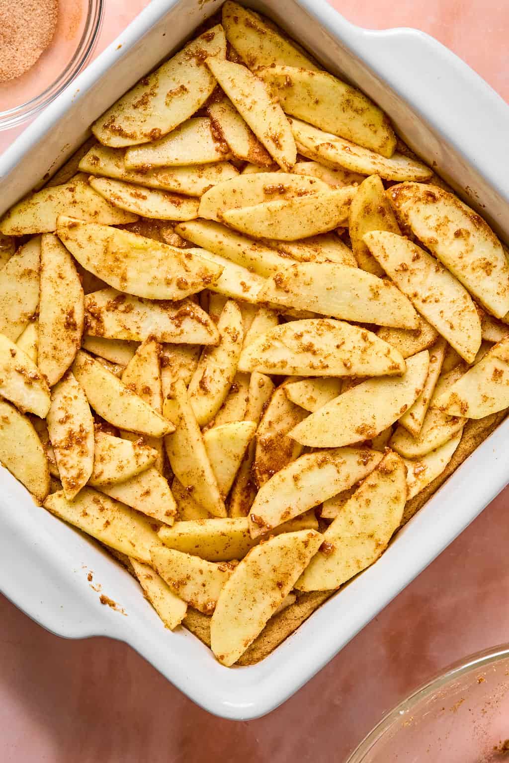 A white baking dish filled with sliced apples coated in cinnamon and sugar, arranged in layers like the filling for apple pie bars, ready to be baked.