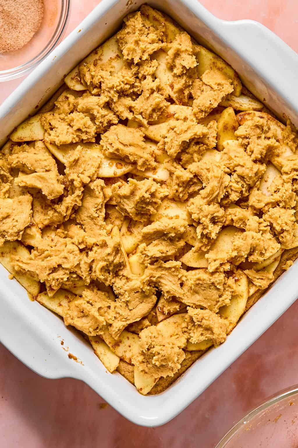 Unbaked apple pie bars with snickerdoodle topping in a white square baking dish, on a pink surface.