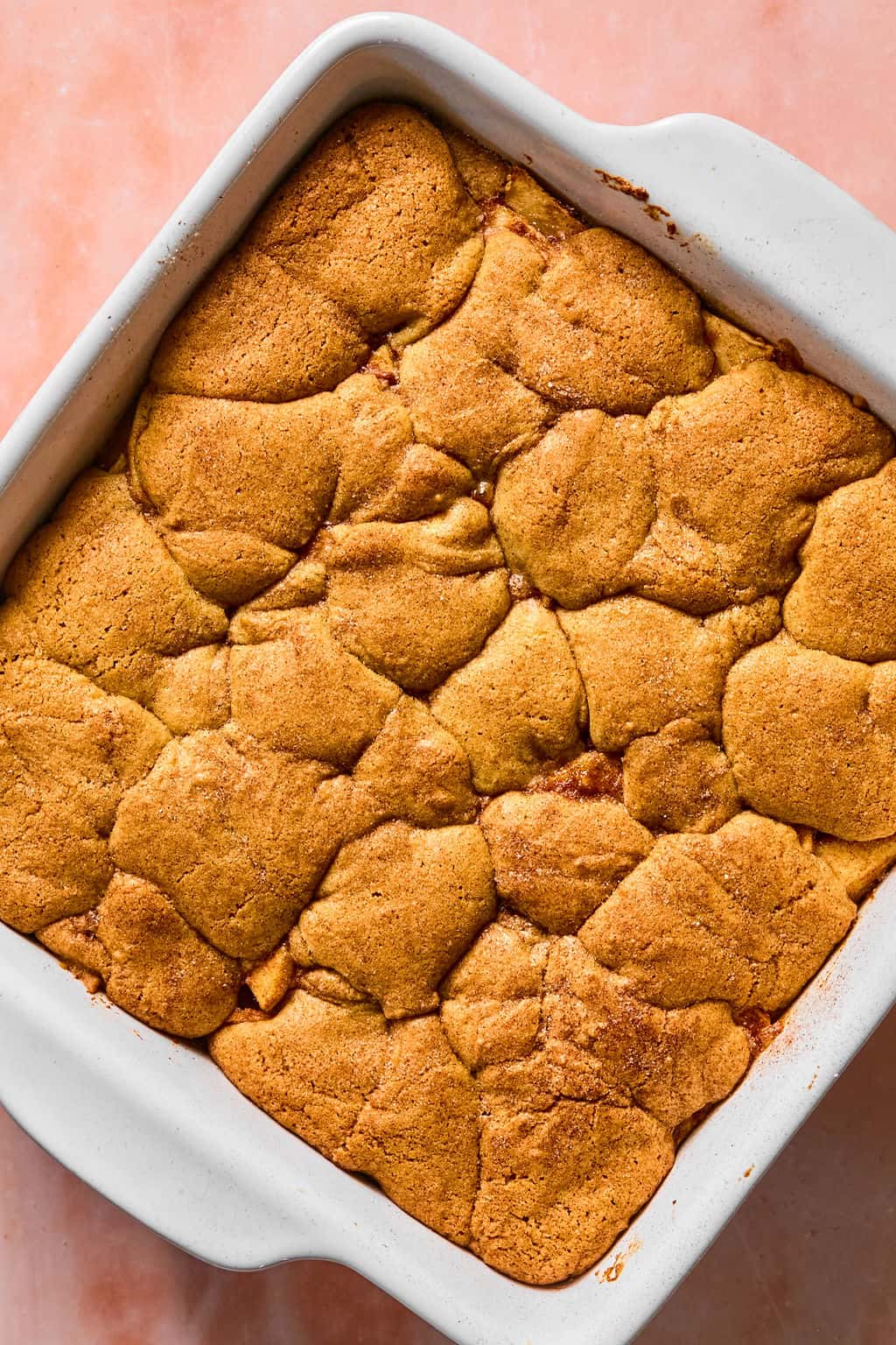 Freshly baked golden-brown apple pie bars with a slightly cracked, textured top sits in a square white baking dish on a pink surface.