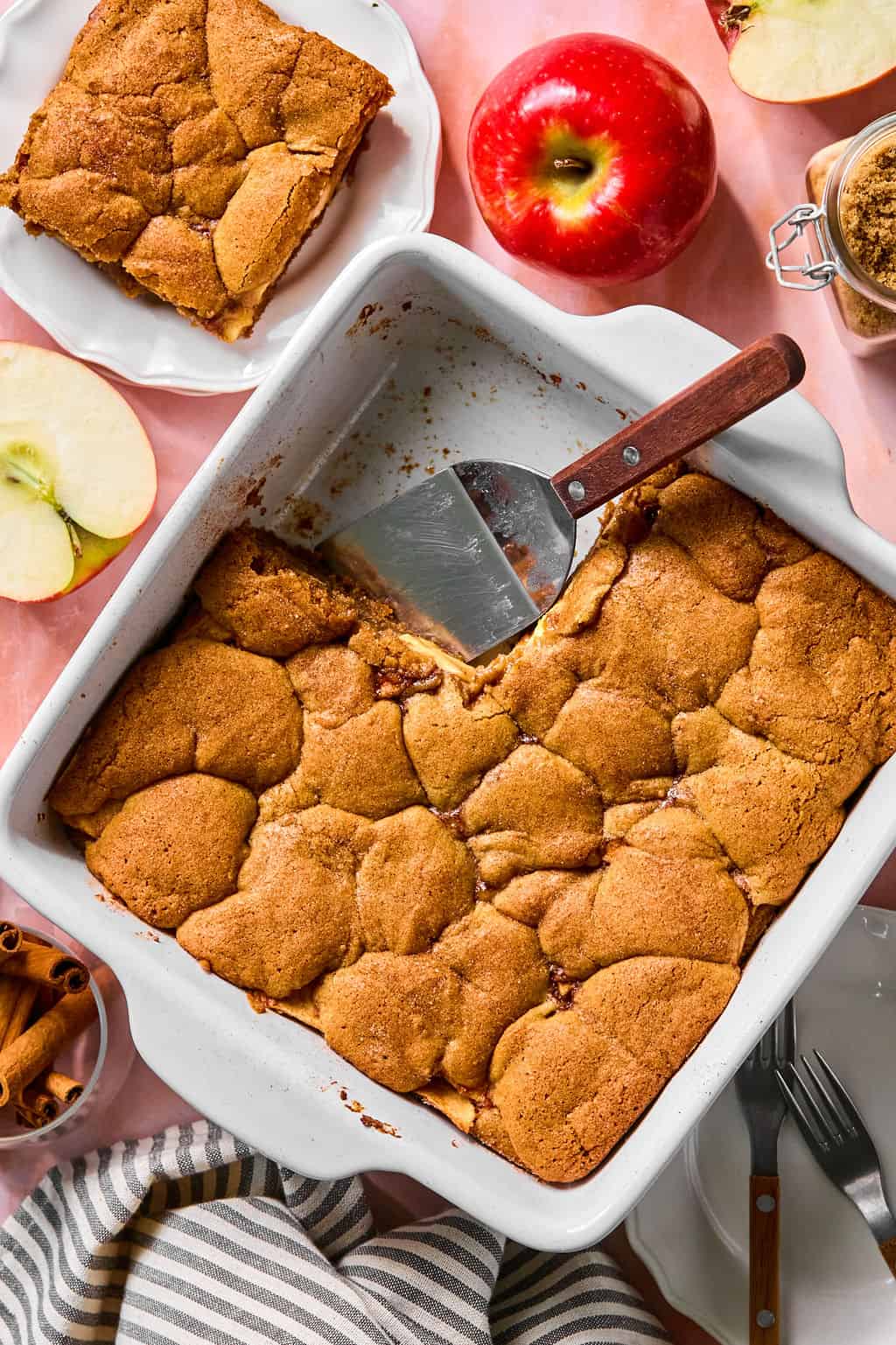 A baking dish of apple pie bars with a serving spoon, a slice on a plate, a red apple, apple slices, a jar of brown sugar, cinnamon sticks, and striped towel on a pink surface.