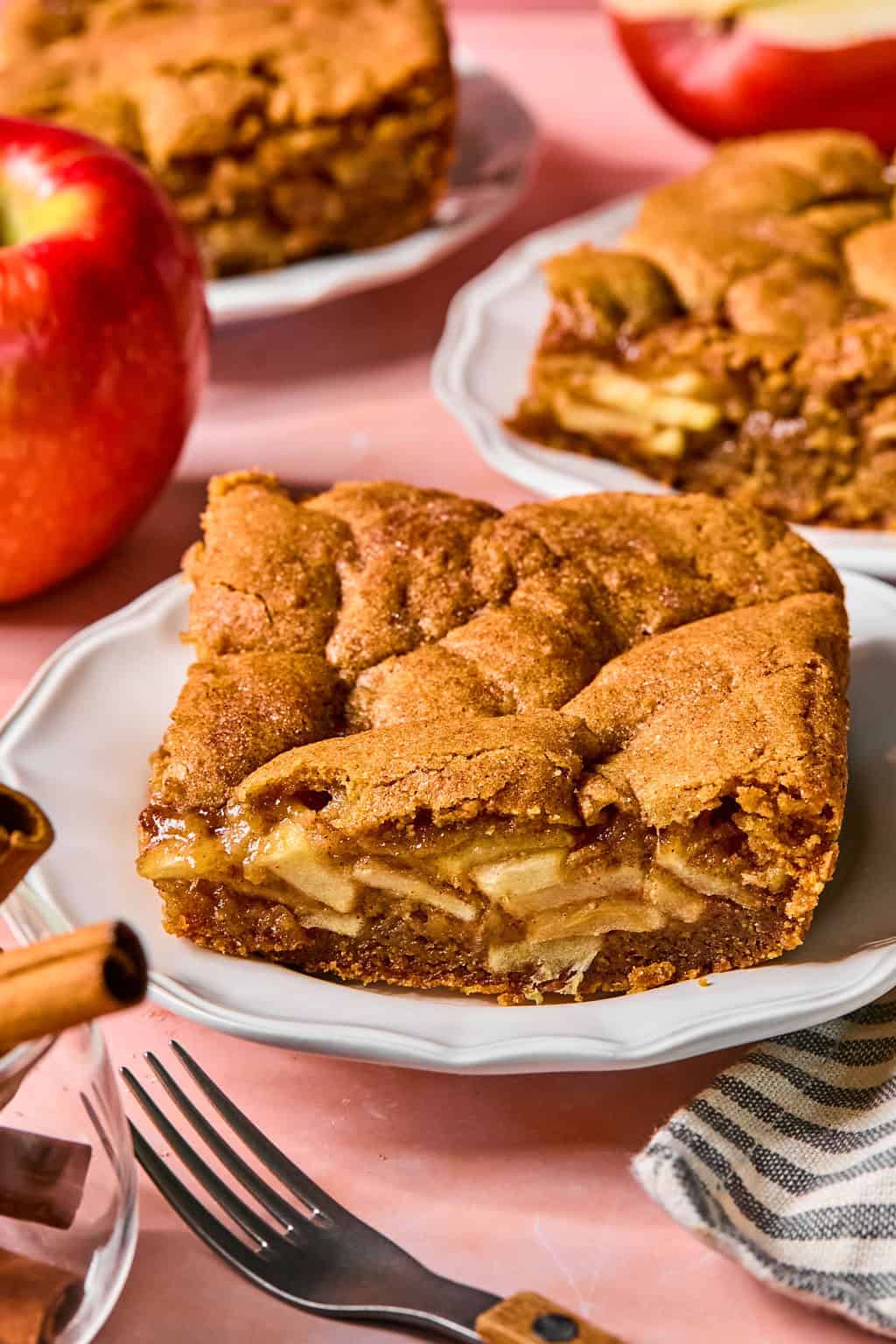 A square slice of apple pie bar with a golden-brown crust, layered with apple slices, sits on a white plate. Whole apples, cinnamon sticks, and more apple pie bars are in the background.