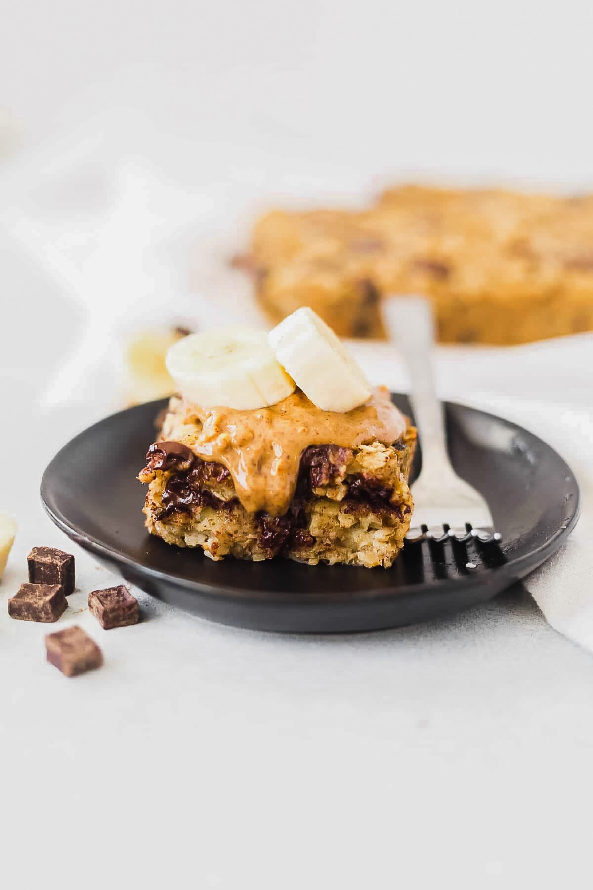 a slice of banana baked oatmeal on a black dessert dish topped with almond butter and banana and served with a fork 