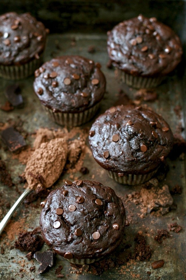 healthy double chocolate zucchini muffins resting on a counter with a spoon full of coco powder and broken pieces of chocolate