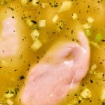 Raw chicken breasts cooking in golden broth with chopped veggies and herbs in a large soup pot.