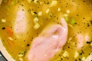 Raw chicken breasts cooking in golden broth with chopped veggies and herbs in a large soup pot.