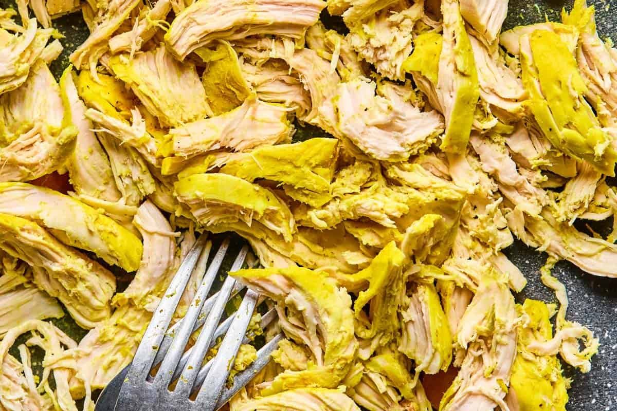 Shredded turmeric-seasoned chicken on a cutting board with a fork, prepared for chicken soup.