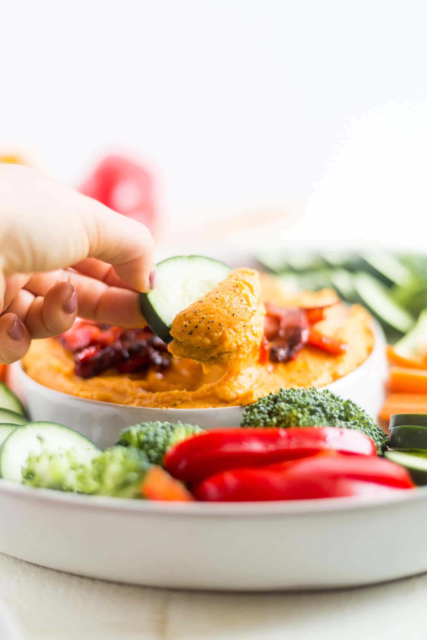 hand dipping into sweet potato hummus with a cucumber slice