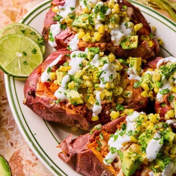 Baked taco stuffed sweet potatoes topped with corn, avocado, cilantro, and a creamy white sauce, served on a white plate with green stripes and lime slices on the side.