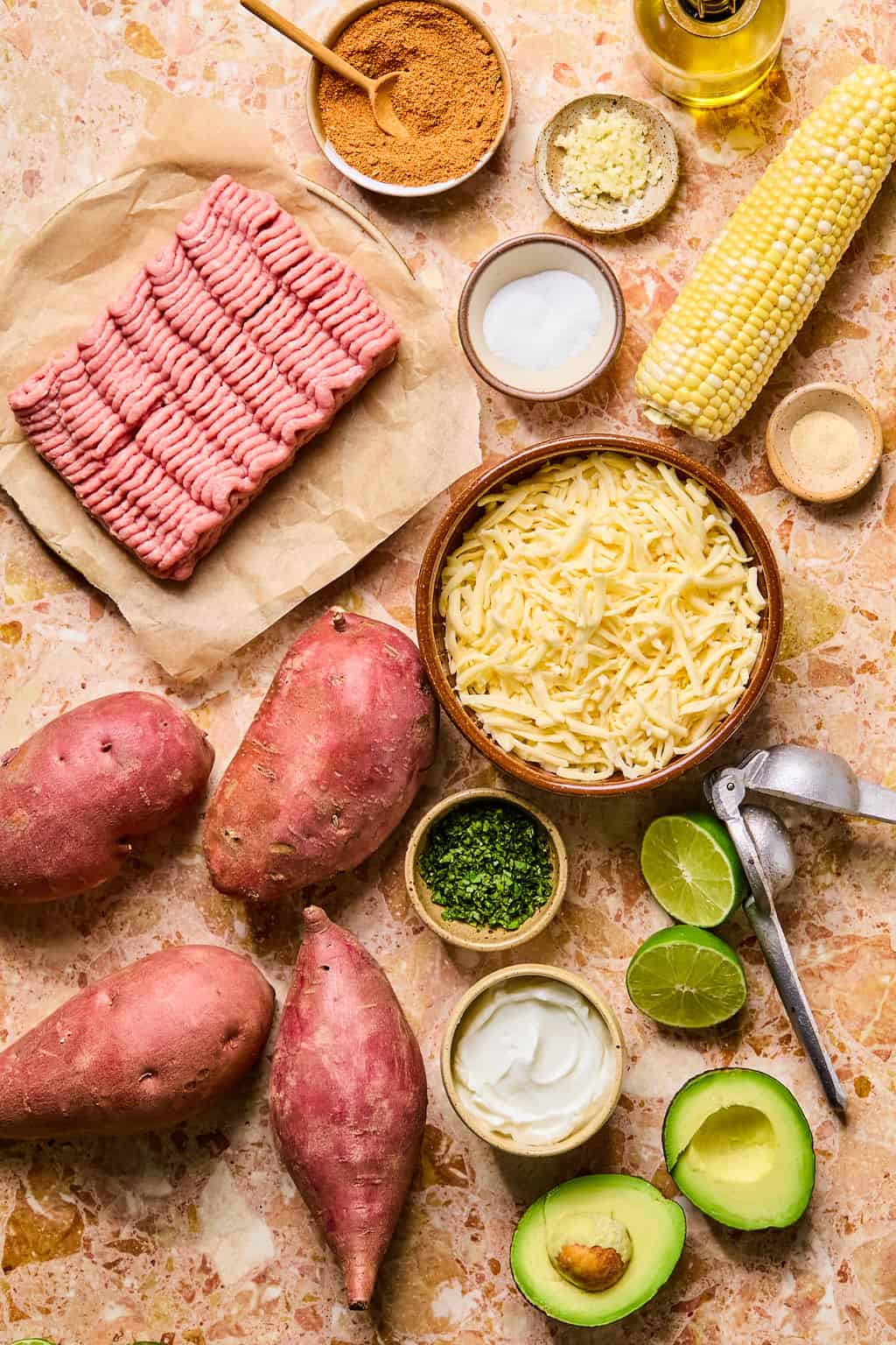Raw ground turkey, shredded cheese, sweet potatoes, corn, avocado, sour cream, lime, chopped herbs, spices, oil, and seasoning are arranged neatly on a marble surface—perfect for creating delicious tacos stuffed sweet potato.