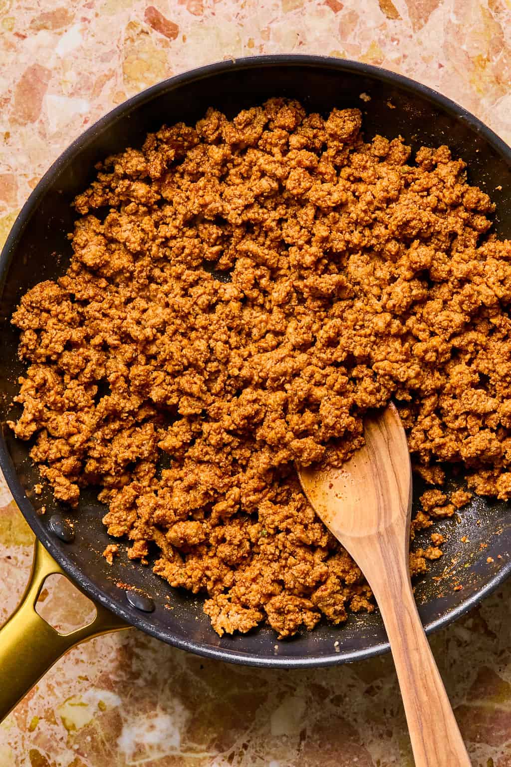 Cooked, seasoned ground taco meat in a black skillet with a wooden spoon resting on the side, ready to be used for delicious tacos or as a hearty filling for stuffed sweet potato, placed on a marbled countertop.
