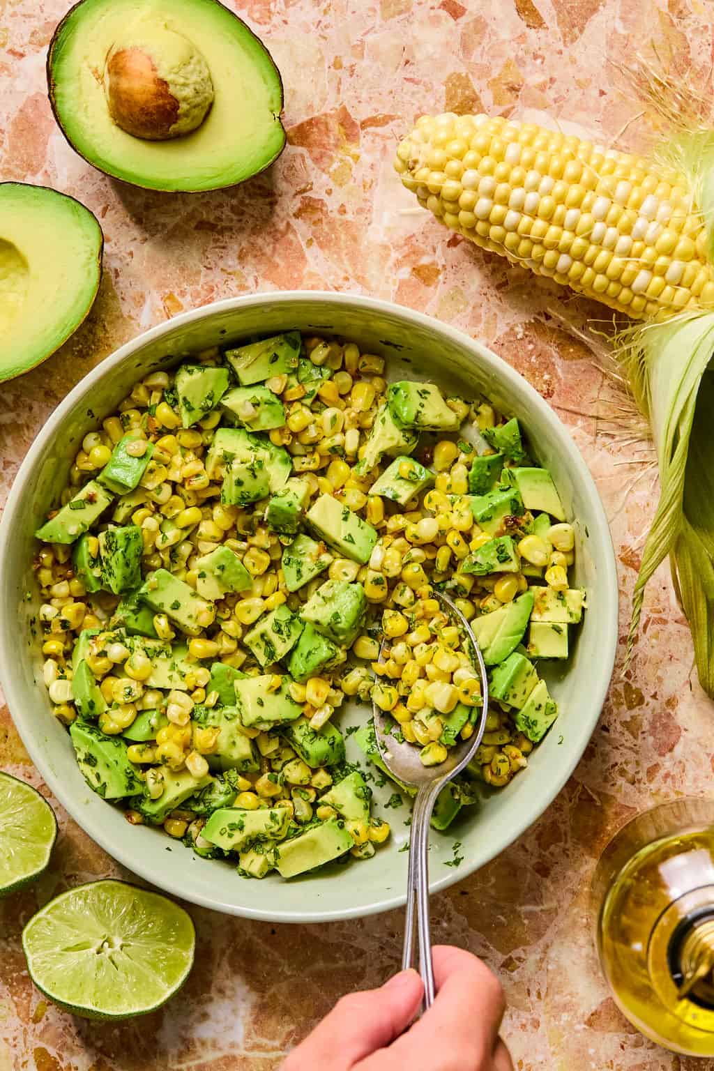 A bowl of corn and avocado salsa with chopped cilantro, surrounded by halved avocados, corn on the cob, lime halves, and a hand holding a spoon, rests on a marble surface—perfect as a vibrant side for tacos or stuffed sweet potato.