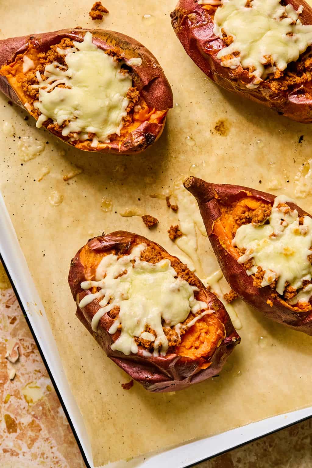 Four tacos stuffed sweet potato halves topped with ground meat and melted cheese, placed on parchment paper on a baking sheet.
