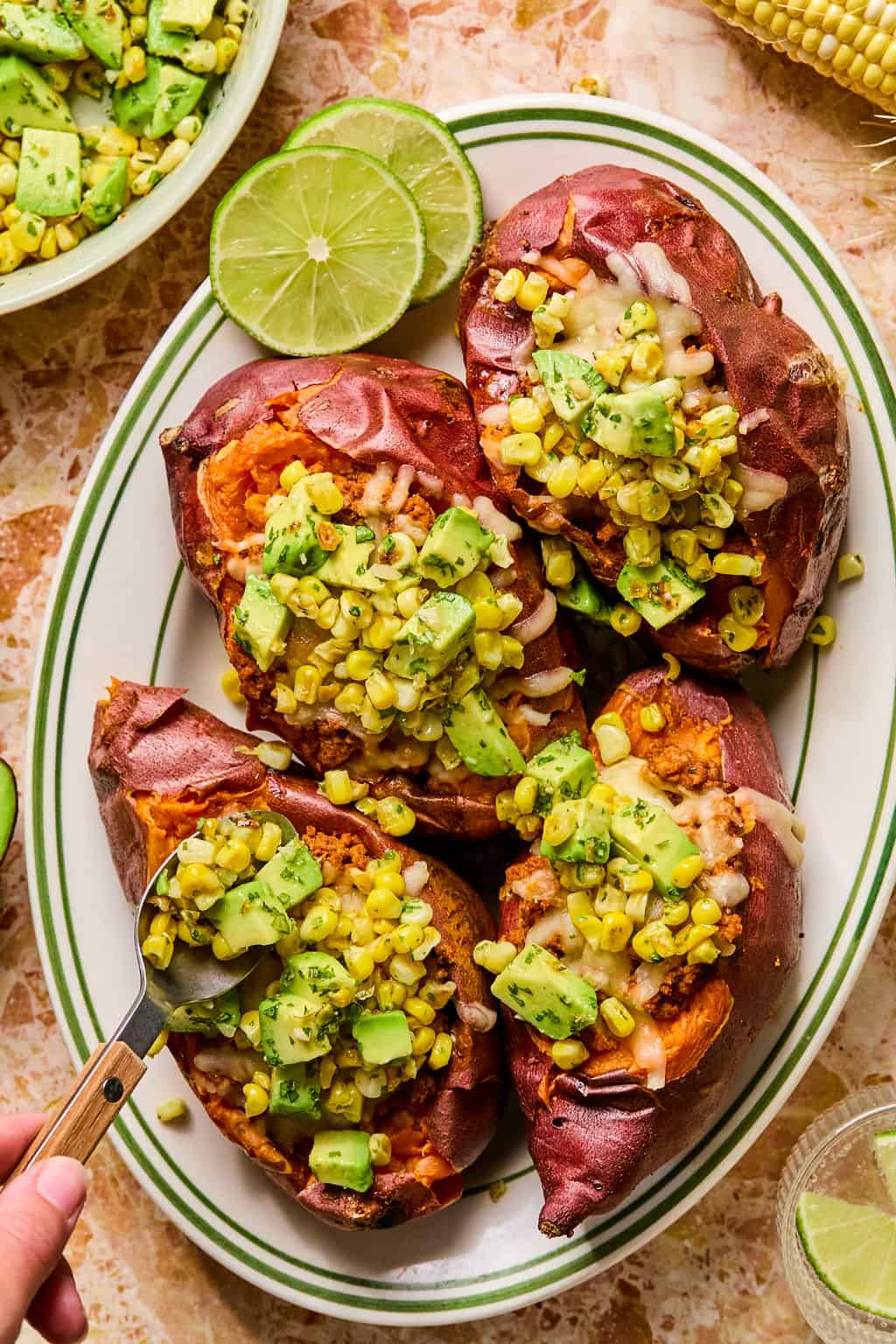 Four tacos stuffed sweet potatoes on a plate, topped with corn, diced avocado, herbs, and melted cheese. A halved lime is on the side, and a hand holds a spoon near one potato. The scene is colorful and appetizing.