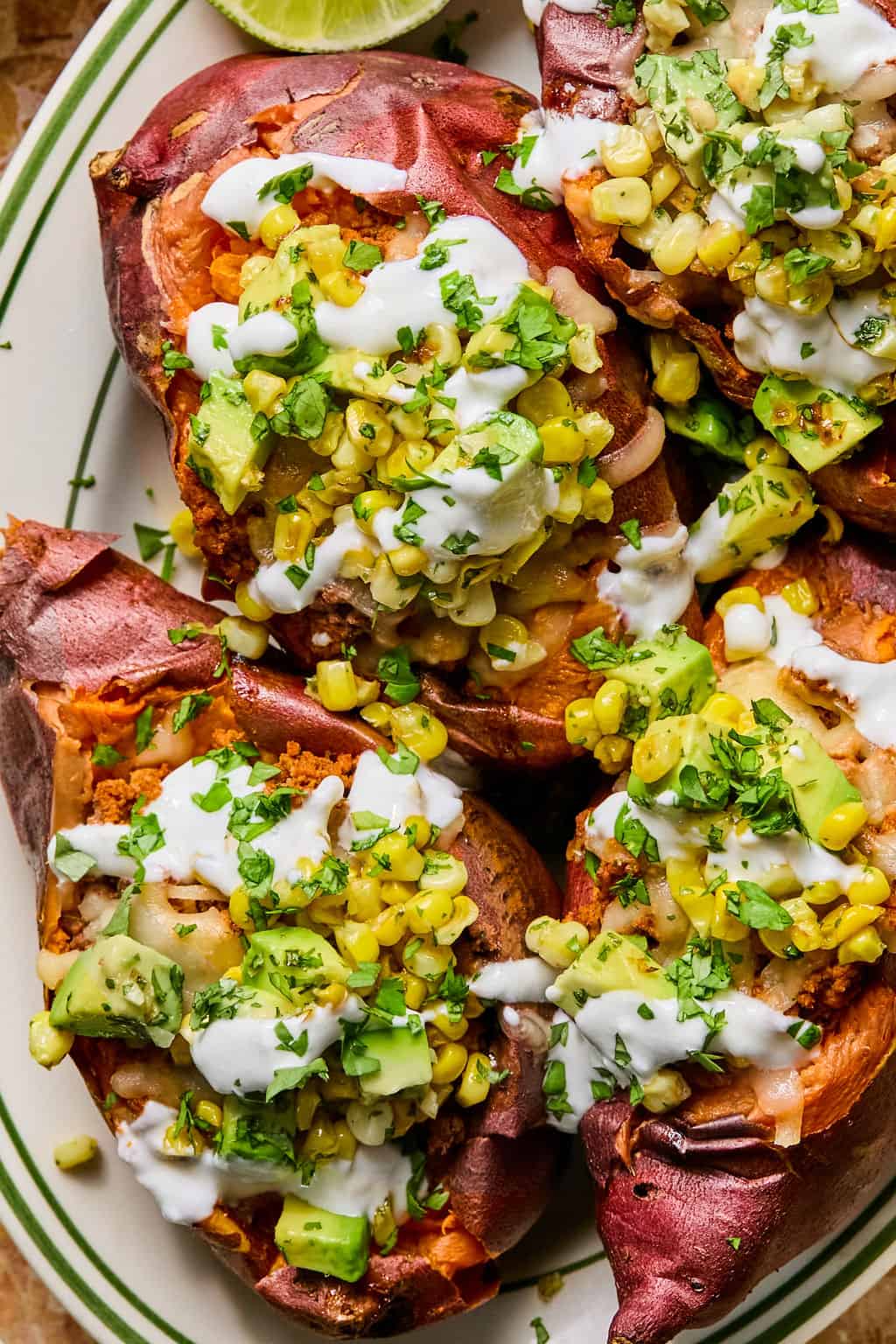 Tacos stuffed sweet potato topped with corn, avocado, onions, fresh herbs, and drizzled with a white creamy sauce, served on a white plate with green stripes.
