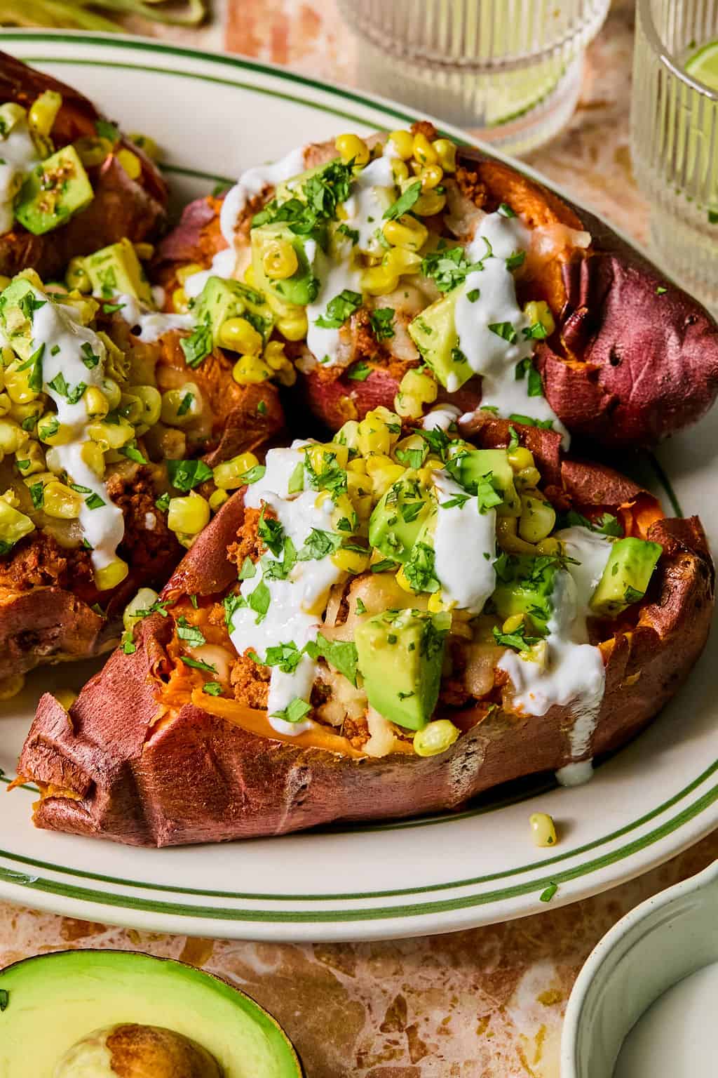 Baked sweet potatoes are transformed into tacos stuffed sweet potato style, topped with avocado, corn, chopped herbs, and a drizzle of creamy white sauce. Served on a white plate with green trim, the dish is complemented by sliced avocado and glasses of water in the background.