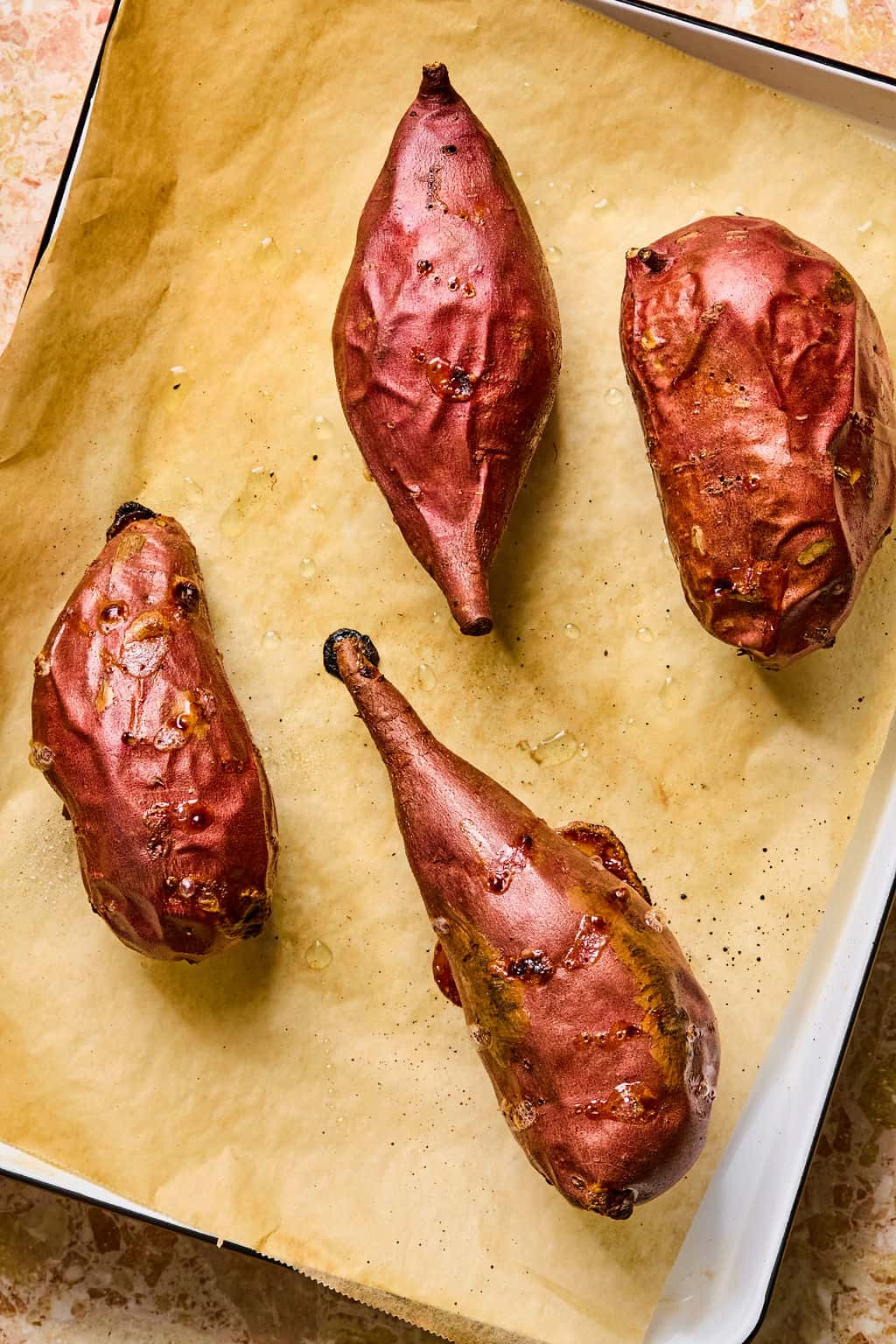 Four roasted sweet potatoes with reddish skins rest on a parchment-lined baking sheet, their slightly wrinkled, caramelized surfaces hinting at rich flavor. Perfectly spaced apart on the tray, these sweet potatoes make an ideal base for tacos stuffed sweet potato creations.