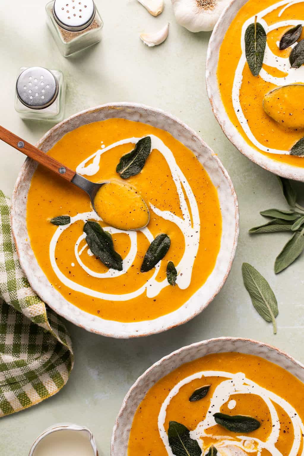 three bowls of butternut squash soup on a counter with salt and pepper and a serving spoon.