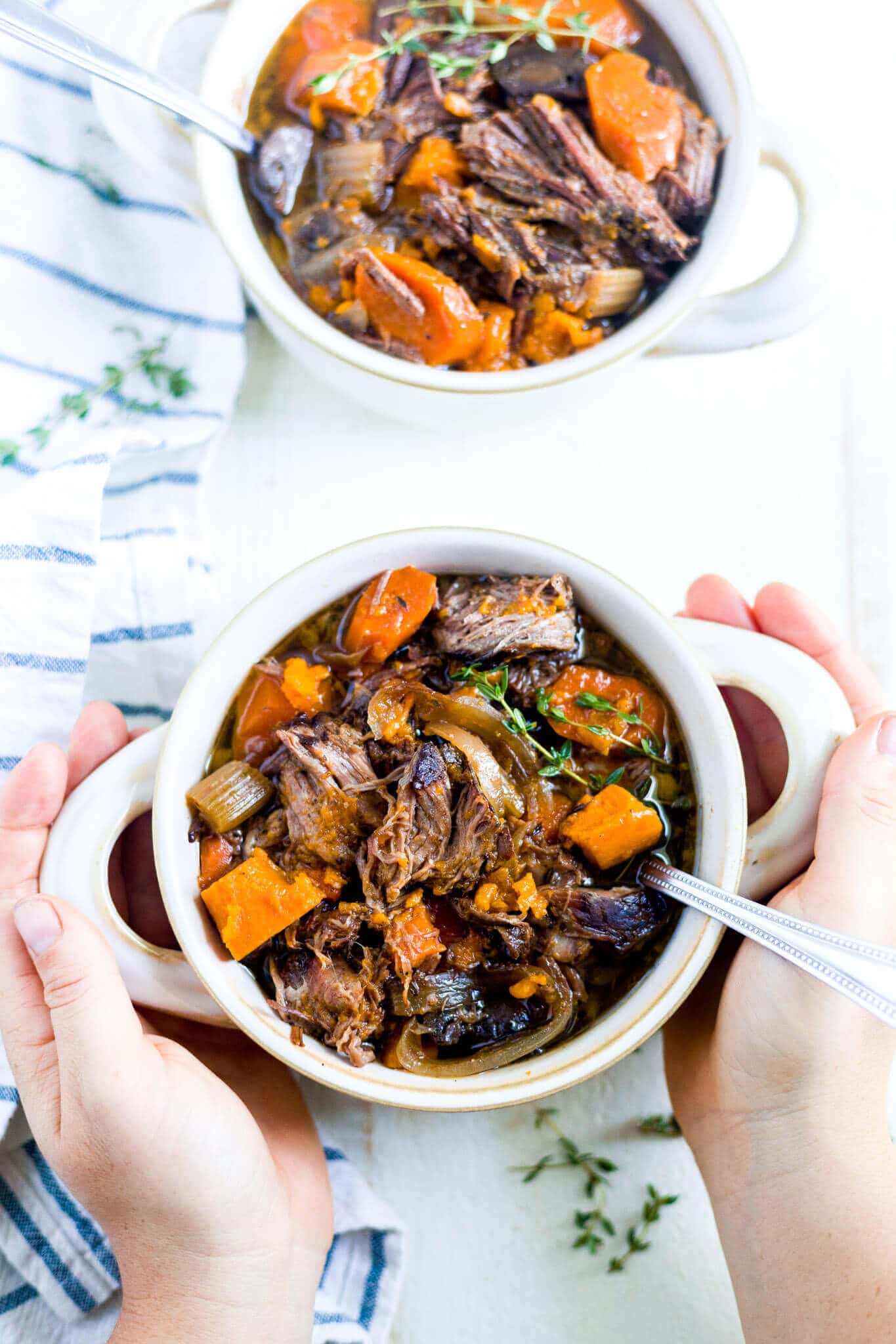 two hands holding a bowl of beef and sweet potato stew with a spoon