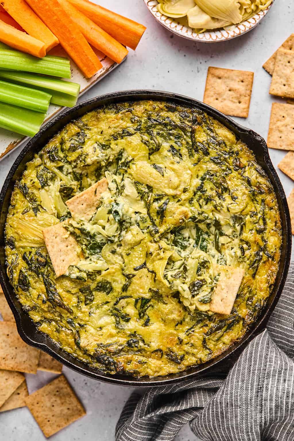 dairy free spinach artichoke dip in a skillet with crackers and veggies on the side.