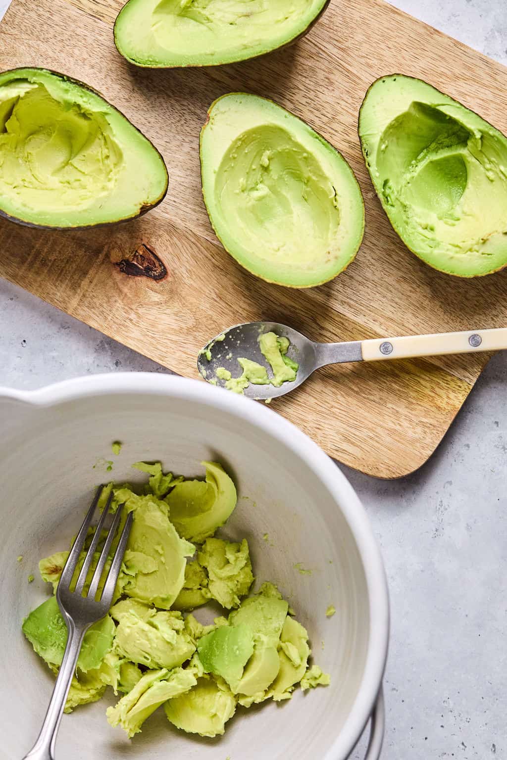 a fork sitting in a white cermaic bowl of avocado next to a wooden cutting board with a spoon resting on it along with avocado halves with avocado scooped out of them