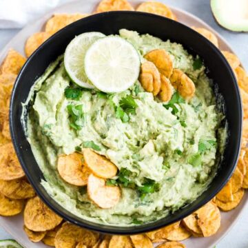 avocado dip in a black bowl with lime, cilantro and plantain chips